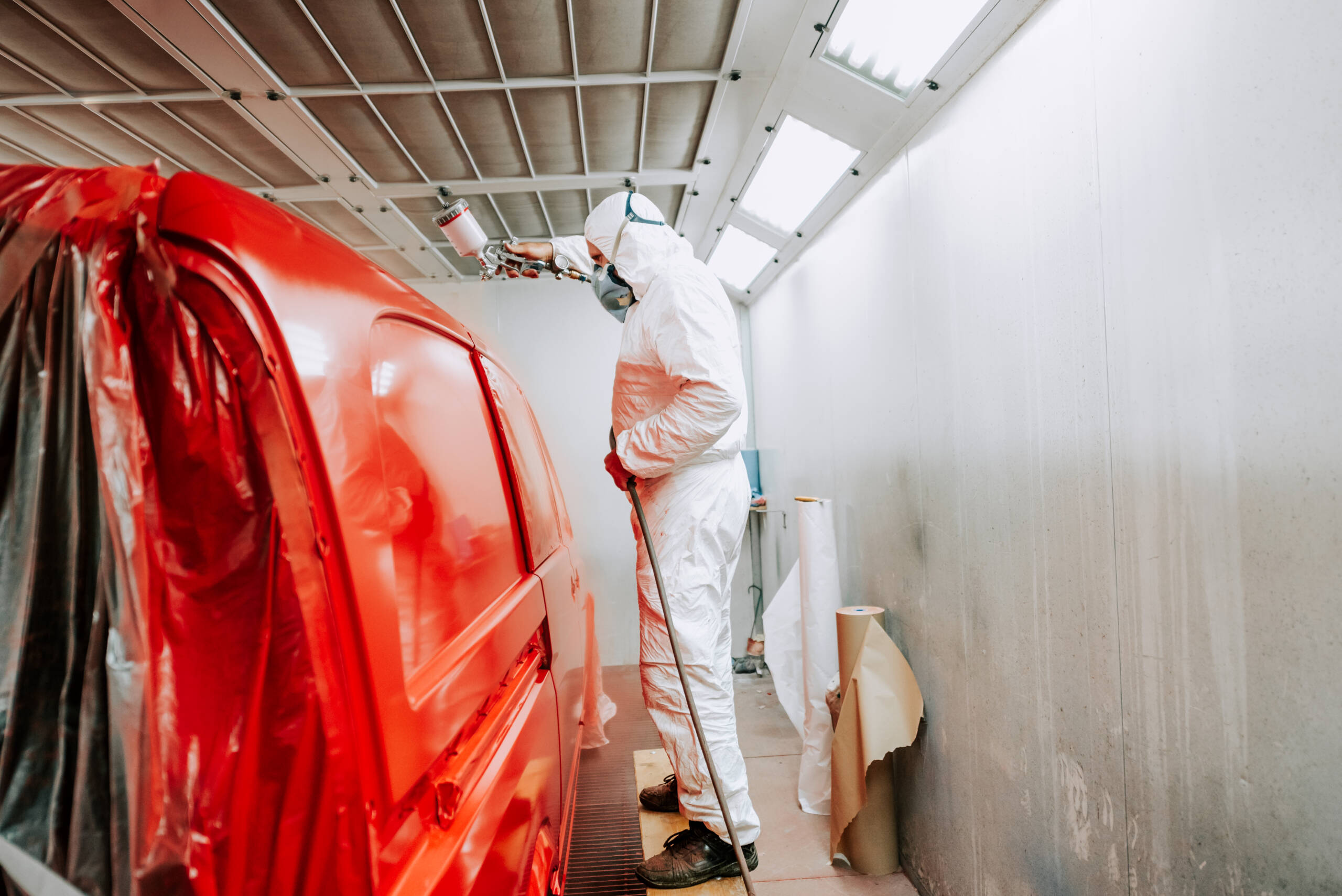 Trabajador pintando un coche rojo en una caja de pintura especial, vestido con un traje blanco y equipo de protección.
